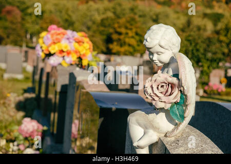 Statue eines Engels hält eine Rose sitzt auf einem Grabstein auf dem Friedhof mit Blumen und Grabsteine im Hintergrund Stockfoto