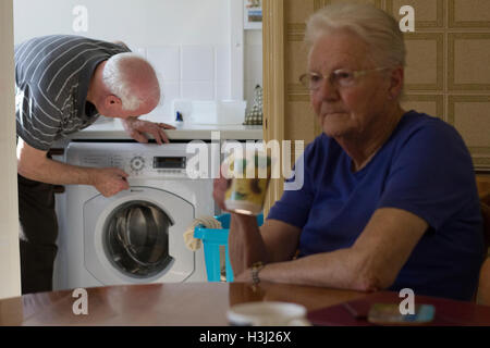 Holmes Chapel, England.14th Mar, 2016. Eine Frau sitzt am Tisch und ein Mann schaut auf eine Waschmaschine. © AimeeHerd Freelance Stockfoto