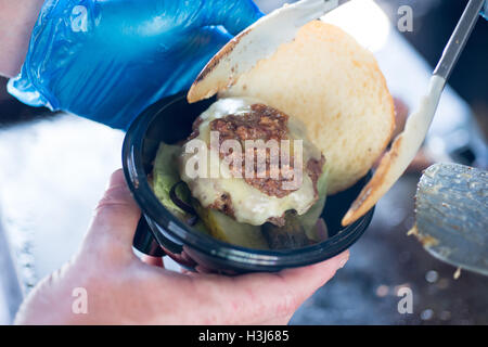 Cheeseburger mit gezogenes Schweinefleisch, platziert auf einem Brötchen mit Gurken in einem schwarzen Kunststoff-Lebensmittel-Container von behandschuhten Händen gehalten Stockfoto