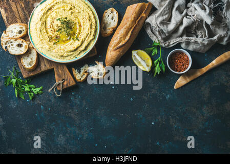 Hausgemachter Hummus Tauchen in vie Keramikschüssel mit frischem Baguette, Zitrone, Kräuter und Gewürze über dunkle blaue Sperrholz Hintergrund, Top blau Stockfoto