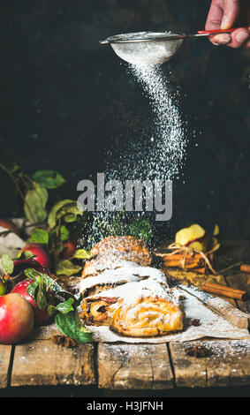 Die Hand des Mannes mit Sieb Beregnung Puderzucker auf Strudel Apfelkuchen mit Zimt und frischen Äpfeln auf rustikalen Holztisch, dunkel Stockfoto