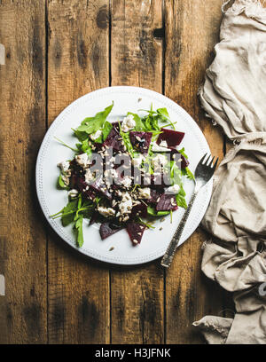 Rucola, rote Beete, Feta-Käse und Sonnenblumenkerne Salat in weißen Keramikplatte über rustikale Holz Hintergrund, Draufsicht, vertikale c Stockfoto