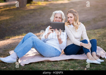 Zwei emotionale Frauen sitzen auf dem Rasen und mit Tablet. Stockfoto