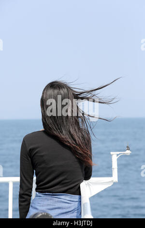 Rückansicht des jungen Asiatin mit wehenden langen Haaren mit Blick auf das Meer vom Deck des Kreuzfahrtschiffes Stockfoto