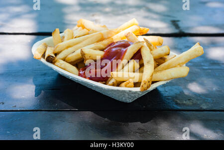 Frische Pommes Frites mit Ketchup in Papier-Container auf Holztisch in teilweise Licht und Schatten im Freien, von der Seite betrachtet. Stockfoto