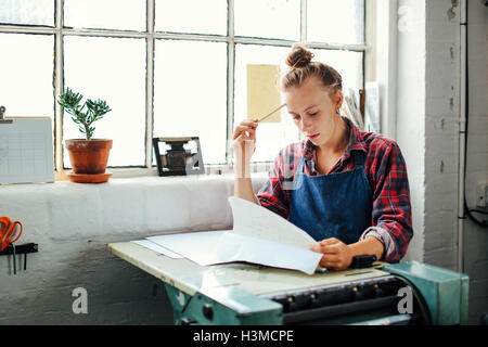 Junge Handwerkerin Fenster, stützte sich auf Buchdruck Maschinen und mit Blick auf Papierkram im Buch-Kunst-studio Stockfoto