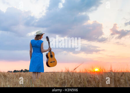 Junge hübsche Frau zu Fuß im Weizenfeld bei Sonnenuntergang mit akustischer Gitarre in der hand, um Inspiration für Country-Songs zu finden Stockfoto