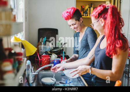 Zwei junge Frauen mit rosa Haaren lachen während bei Küchenspüle Abwasch Stockfoto