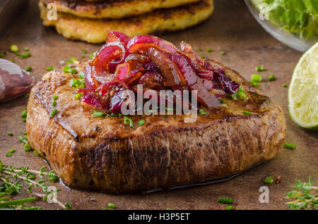 Leckeres Rindersteak mit Pfeffer, Knoblauch und karamellisierten Zwiebeln, Kartoffelpuffer hinter Stockfoto