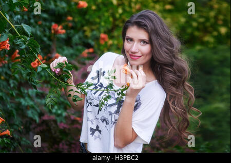 Schöne junge Frau mit langen lockigen Haaren posiert in der Nähe von Blumen in einem Garten Stockfoto