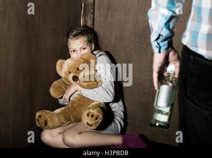 Trauriges kleines Mädchen sitzt an der Wand in Verzweiflung. In seinen Händen hält er ein Teddybär. betrunkener Mann droht. Stockfoto