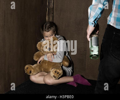 Trauriges kleines Mädchen sitzt an der Wand in Verzweiflung. In seinen Händen hält er ein Teddybär. betrunkener Mann droht. Stockfoto