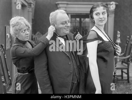 1910ER JAHRE 1920ER JAHRE ÄLTEREN LÄCHELND MANN ZERRTE AN JUNGE FRAU KLEID, WÄHREND DIE FRAU AUF DES MANNES OHR STUMMFILM NOCH SCHLEPPER Stockfoto