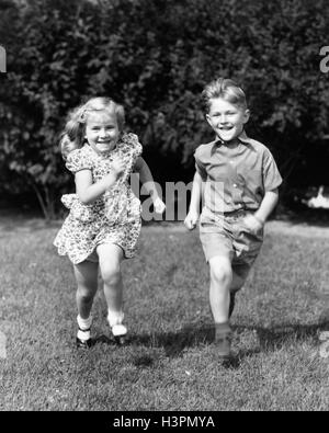 1930S 1940S GRASS LÄCHELNDEN JUNGEN UND MÄDCHEN LAUFEN IM SOMMER GARTEN BLICK IN DIE KAMERA Stockfoto