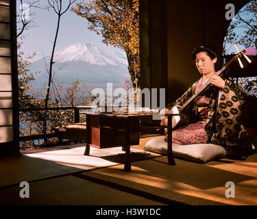 1960ER JAHRE JAPANERIN MUSIKER SPIELT EIN SHAMISEN SAITENINSTRUMENT SITZEN IN EINEM TRADITIONELLEN HAUS IN DER NÄHE VON MT. FUJI, JAPAN Stockfoto