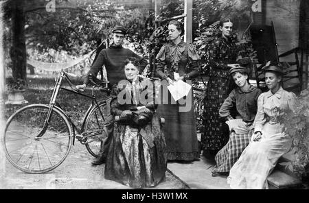 1890ER JAHRE GRUPPE PORTRAIT FÜNF FRAUEN EINEN MANN MIT FAHRRAD AUF DER VERANDA DES HAUSES, BLICK IN DIE KAMERA ZU POSIEREN Stockfoto