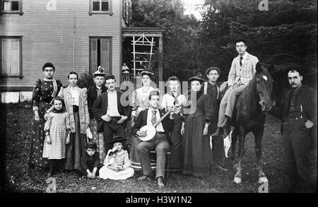 1890ER JAHRE GRUPPE PORTRAIT FAMILIE BLICK AUF KAMERA POSIERT AUF DER WIESE VOR HAUS MANN IN MITTE HOLDING BANJO MANN SITZT AUF PFERD Stockfoto