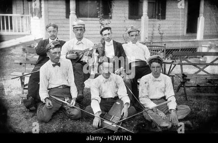 1900ER JAHRE GRUPPE PORTRAIT MÄNNER POSIEREN MIT MUSIKINSTRUMENTEN MANDOLINE AUTOHARP UND ANGELRUTEN SITZEN AUF DER WIESE VOR HAUS Stockfoto