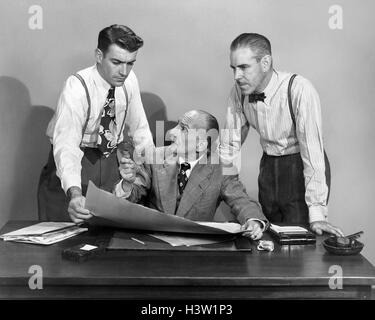 1950ER JAHREN DREI GESCHÄFTSLEUTE IM BÜRO STRATEGIE KONFERENZ ZWEI STEHEN EINER SITZEN, BLICK AUF PAPIERE UND PLÄNE Stockfoto