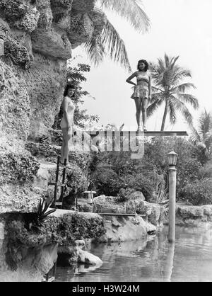 1930ER JAHREN 1940ER JAHREN 2 FRAUEN POSIEREN POOL SEITE TRAGEN BADEANZÜGE TROPISCHEN LAGE PALMEN Stockfoto