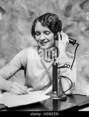 1920ER JAHRE LÄCHELNDE FRAU SITZT AM SCHREIBTISCH, SCHREIBEN UND SPRECHEN AUF CANDLESTICK-TELEFON Stockfoto