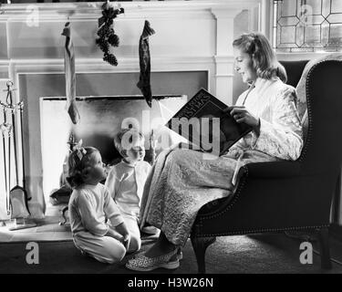 1940S 1950S MUTTER LESUNG WEIHNACHTEN GESCHICHTE FÜR JUNGE MÄDCHEN SITZEN VON KAMIN HUNG MIT STRÜMPFEN Stockfoto