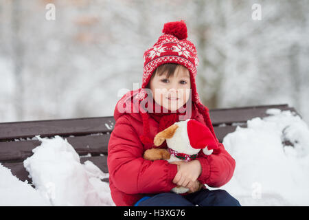 Entzückende kleine Kind, junge, spielen in einem verschneiten Park holding Teddybär, sitzen auf Bank, Winter Stockfoto