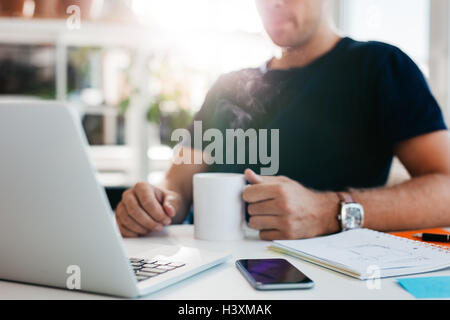 Schuss der Geschäftsmann sitzt an seinem Schreibtisch hält eine Tasse Kaffee beschnitten. Laptop, Handy und Tagebuch auf dem Tisch. Stockfoto