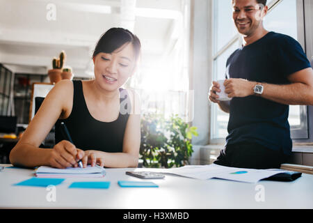 Junge asiatische Frau Schreiben von Notizen mit männlicher Kollege mit Tasse Kaffee bereit. Kollegen zusammen arbeiten im Büro Stockfoto