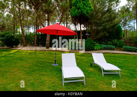 Zwei weiße Stoff-Lounge-Sesseln und einem roten Sonnenschirm auf einer Wiese im Garten Stockfoto