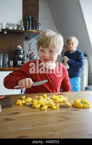 Glücklich behinderten jungen hacken gelbe Paprika mit Bruder im Hintergrund in Küche Stockfoto