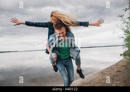 Glücklicher Mann Huckepack Freundin am Seeufer gegen Himmel Stockfoto
