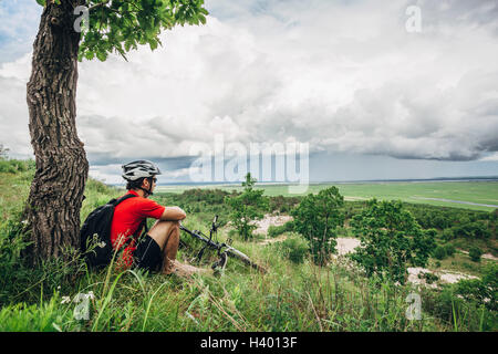 Müde Mountainbiker unter Baum auf Hügel gegen bewölktem Himmel ruht Stockfoto