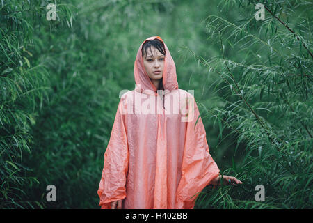 Junge Frau trägt Regenmantel stehen inmitten von Pflanzen während der Regenzeit Stockfoto