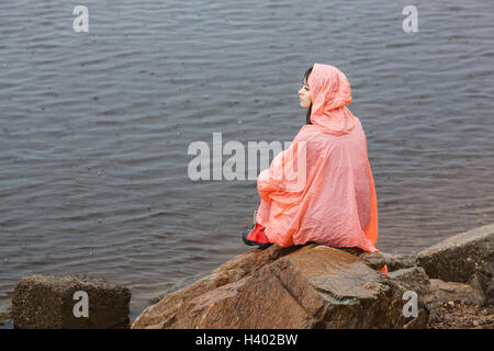 Nachdenkliche Frau mit Regenmantel während der Regenzeit auf Felsen am Ufer sitzen Stockfoto