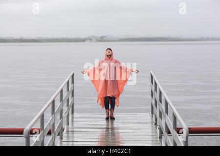 Frau trägt Regenmantel genießen Regenzeit auf Steg stehend Stockfoto