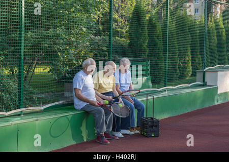 Senioren Freunde mit Tennisschläger sitzen gegen Zaun am Hof Stockfoto