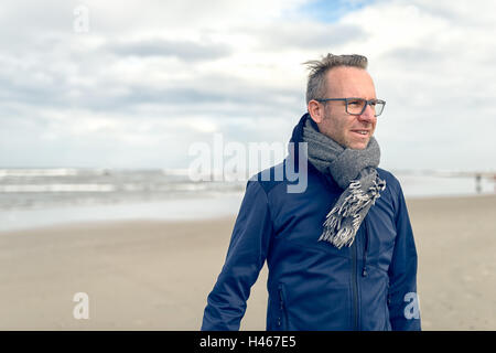 Gereizter Mann mittleren Alters tragen Brillen und einen Schal aus Wolle Stand an einem einsamen Strand im Herbst an einem bewölkten Tag mit c Stockfoto