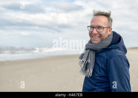 Lächelnder Mann mittleren Alters tragen Brillen und einen Schal aus Wolle Stand an einem einsamen Strand im Herbst an einem bewölkten Tag mit cop Stockfoto