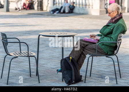 Ein senior Mann sitzt alleine tun ein Kreuzworträtsel in den Hof des Somerset House, London. Stockfoto