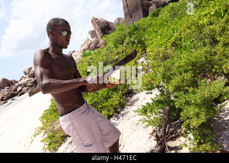 Die Seychellen, Strand, junger Mann öffnet Kokosnuss, Stockfoto