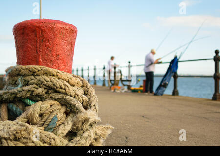 Roter Stein Liegeplatz Poller mit einem Seil gebunden um ihn herum auf Torquay Pier wo unbekannte Fischer ihre Angelschnüre vorzubereiten Stockfoto