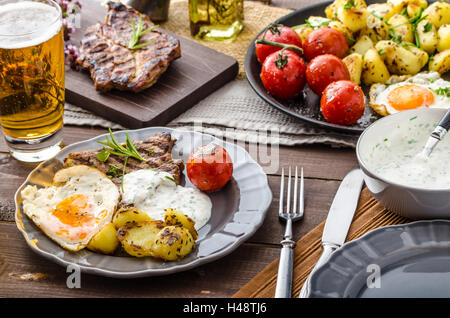 Gegrilltes Schweinefleisch mit Bier, Knoblauch-Dip, gerösteten Kartoffeln, Tomaten und Spiegelei Stockfoto