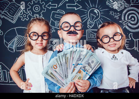 Kleiner Junge in Gläsern und Schnurrbart auf dunklem Hintergrund mit einem Muster. In der hand hält er Banknoten. Zwei Mädchen stehen die Seiten Stockfoto