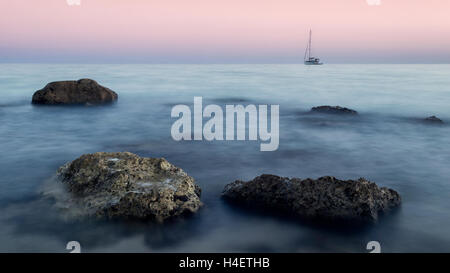 Einsame Boot am Horizont über einem felsigen Ufer im Sonnenaufgang Stockfoto