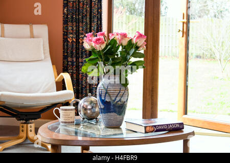 Ein Lifestyle-Bild ein Couchtisch mit Rosen, Teekanne, Becher & Buch in eine durchschnittliche UK nach Hause mit einem Garten Outlook. Stockfoto