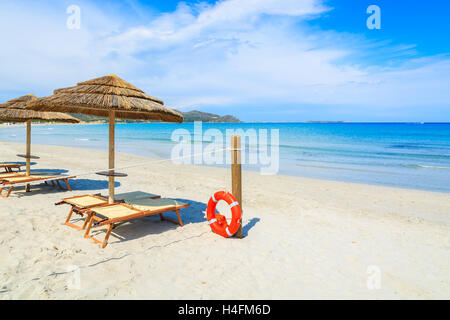 Liegestühle mit Sonnenschirmen und roten Rettungsring auf eine Holzstange am weißen Sandstrand, Porto Giunco Bay, Insel Sardinien, Italien Stockfoto