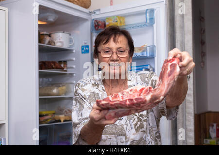 Ältere Frau mit rohe Schweinerippchen vor den offenen Kühlschrank steht in der Küche. Stockfoto