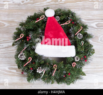 Immergrüne Adventskranz mit Weihnachtsmann Mütze, Lichter, Tannenzapfen und Zuckerstangen auf weißem Holz. Stockfoto