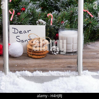 Blick aus einem Stapel von Cookies, Glas Milch und Brief an den Weihnachtsmann mit immergrünen Dekorationen im Hintergrund durch verschneite Fenster. Stockfoto